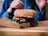 A close-up view of a multigrain sandwich with sesame seeds, filled with fresh vegetables including tomato slices and leafy greens. The sandwich is placed on a wooden board with hands partially visible in the background, suggesting a meal setting.