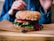 A close-up view of a multigrain sandwich with sesame seeds, filled with fresh vegetables including tomato slices and leafy greens. The sandwich is placed on a wooden board with hands partially visible in the background, suggesting a meal setting.