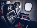 Two leather airplane seats are situated in a row by the windows, with red seatbelts fastened across them. The plane’s cabin lighting casts a soft glow on the seats, creating a sense of calm and order.
