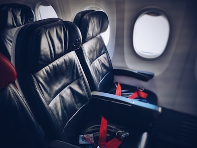 Two leather airplane seats are situated in a row by the windows, with red seatbelts fastened across them. The plane’s cabin lighting casts a soft glow on the seats, creating a sense of calm and order.