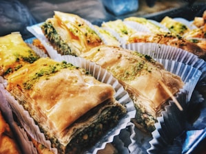 Slices of baklava with a shiny, golden-brown surface and layers of flaky pastry filled with a green, nutty mixture. Each piece is placed in a white paper baking cup, arranged closely together.