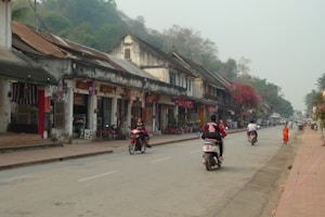 A quaint street scene with a row of rustic buildings lining the road. People are riding scooters along the street, while a young person in orange robes walks on the sidewalk. The buildings have a historical look, with weathered textures and flags hanging outside. Lush greenery is visible in the background, giving a serene and culturally rich atmosphere.