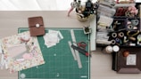 A crafting workspace featuring a green cutting mat with rulers, scissors, a crafting knife, and floral scrapbook paper. On the left is a closed brown notebook, while the right side holds a variety of stationery items, including tape, stamps, pens, and an organized tray with compartments for various crafting materials. There is also a small potted plant adding a touch of greenery.