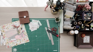 A crafting workspace featuring a green cutting mat with rulers, scissors, a crafting knife, and floral scrapbook paper. On the left is a closed brown notebook, while the right side holds a variety of stationery items, including tape, stamps, pens, and an organized tray with compartments for various crafting materials. There is also a small potted plant adding a touch of greenery.
