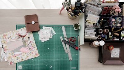 A crafting workspace featuring a green cutting mat with rulers, scissors, a crafting knife, and floral scrapbook paper. On the left is a closed brown notebook, while the right side holds a variety of stationery items, including tape, stamps, pens, and an organized tray with compartments for various crafting materials. There is also a small potted plant adding a touch of greenery.