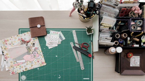 A crafting workspace featuring a green cutting mat with rulers, scissors, a crafting knife, and floral scrapbook paper. On the left is a closed brown notebook, while the right side holds a variety of stationery items, including tape, stamps, pens, and an organized tray with compartments for various crafting materials. There is also a small potted plant adding a touch of greenery.
