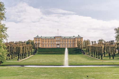 A large, elegant manor house with symmetrical architecture is set amidst sprawling manicured lawns. A prominent water fountain jets upwards in the foreground, creating a focal point. The landscape is dotted with neatly trimmed trees lining the path leading to the house.