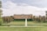 A large, elegant manor house with symmetrical architecture is set amidst sprawling manicured lawns. A prominent water fountain jets upwards in the foreground, creating a focal point. The landscape is dotted with neatly trimmed trees lining the path leading to the house.