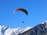 A person is paragliding with a colorful parachute against a clear blue sky. Below, snow-capped mountains create a stunning backdrop, suggesting a high-altitude adventure.