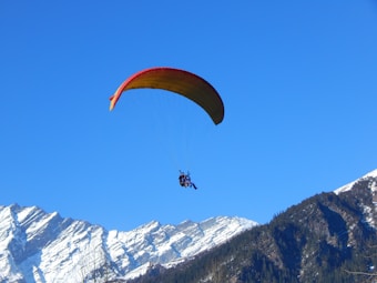 A person is paragliding with a colorful parachute against a clear blue sky. Below, snow-capped mountains create a stunning backdrop, suggesting a high-altitude adventure.