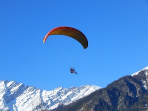 A person is paragliding with a colorful parachute against a clear blue sky. Below, snow-capped mountains create a stunning backdrop, suggesting a high-altitude adventure.