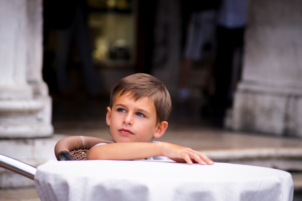 menino olhando para os lados enquanto sentado na cadeira marrom ao ar livre