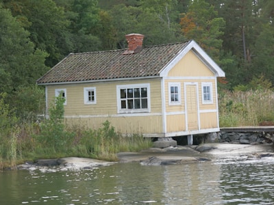 A quaint, small yellow wooden cottage with a chimney is situated on the edge of a serene body of water. The building has white trim and old, weathered roof tiles. Surrounding the cottage is dense, lush greenery, including trees and shrubs, creating a tranquil and secluded atmosphere.