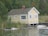 A quaint, small yellow wooden cottage with a chimney is situated on the edge of a serene body of water. The building has white trim and old, weathered roof tiles. Surrounding the cottage is dense, lush greenery, including trees and shrubs, creating a tranquil and secluded atmosphere.