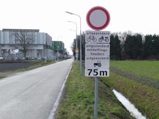 A rural road extends into the distance, bordered by grassy fields and a drainage ditch. A large signpost is visible, featuring a red circle indicating a traffic restriction, with additional signs below showing exceptions for bicycles, mopeds, permit holders, and tractors. The background includes an industrial-looking building and trees lining the road.