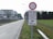 A rural road extends into the distance, bordered by grassy fields and a drainage ditch. A large signpost is visible, featuring a red circle indicating a traffic restriction, with additional signs below showing exceptions for bicycles, mopeds, permit holders, and tractors. The background includes an industrial-looking building and trees lining the road.