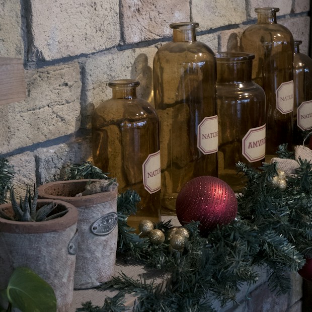 Several amber-colored glass bottles with labels reading 'Nature' and 'Amvrus' are arranged against a brick wall. They are surrounded by green pine garlands and decorative elements, including a red Christmas ornament and small golden balls. There are also two rustic pots containing succulents.