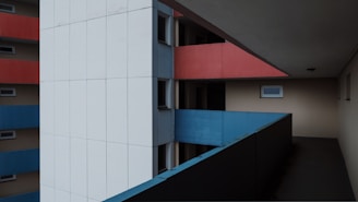A modern architectural design featuring a multi-level building with geometric lines and contrasting colors. The facade is composed of white, red, and blue panels, creating a striking visual. Multiple windows punctuate the surface and a narrow, shadowy corridor extends inward.