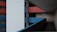 A modern architectural design featuring a multi-level building with geometric lines and contrasting colors. The facade is composed of white, red, and blue panels, creating a striking visual. Multiple windows punctuate the surface and a narrow, shadowy corridor extends inward.