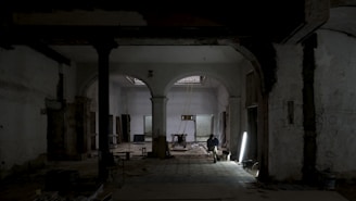 A dimly lit, partially renovated interior with arched doorways and exposed structural elements. A single person sits on a chair near a bright light source on the floor, surrounded by construction materials.