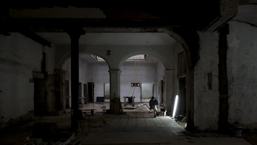 A dimly lit, partially renovated interior with arched doorways and exposed structural elements. A single person sits on a chair near a bright light source on the floor, surrounded by construction materials.