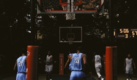 A group of basketball players are engaged in a game on an outdoor court. The scoreboard indicates a close match with only a few minutes remaining. The players are wearing jerseys with numbers, and a basketball hoop is clearly visible. Tall trees surround the court, giving it a secluded and intense atmosphere.