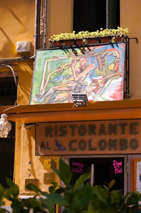 A colorful abstract painting is displayed above the entrance of a building with a sign that reads 'Ristorante al Colombo'. The exterior features a warm yellow hue, and potted plants line the top edge of the artwork. Below, a classic lamp is mounted on the wall, and some greenery is visible in the foreground.