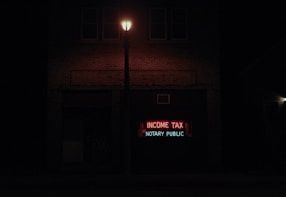 A dimly lit street scene at night features a brick building with a neon sign displaying the words 'INCOME TAX' in red and 'NOTARY PUBLIC' in green. A streetlight illuminates the space immediately around it, creating a contrast with the dark surroundings.