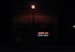 A dimly lit street scene at night features a brick building with a neon sign displaying the words 'INCOME TAX' in red and 'NOTARY PUBLIC' in green. A streetlight illuminates the space immediately around it, creating a contrast with the dark surroundings.