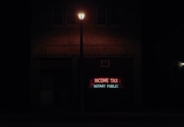 A dimly lit street scene at night features a brick building with a neon sign displaying the words 'INCOME TAX' in red and 'NOTARY PUBLIC' in green. A streetlight illuminates the space immediately around it, creating a contrast with the dark surroundings.