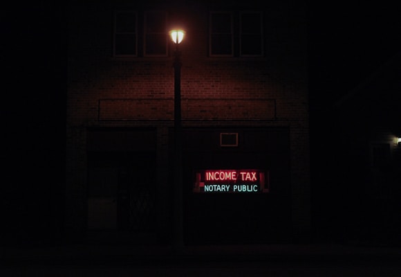 A dimly lit street scene at night features a brick building with a neon sign displaying the words 'INCOME TAX' in red and 'NOTARY PUBLIC' in green. A streetlight illuminates the space immediately around it, creating a contrast with the dark surroundings.