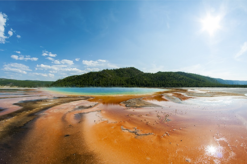 A striking landscape features a vibrant hot spring with orange, yellow, and blue hues in the foreground. Lush green wooded hills and a clear blue sky with scattered clouds provide a stunning backdrop, with the sun shining brightly.