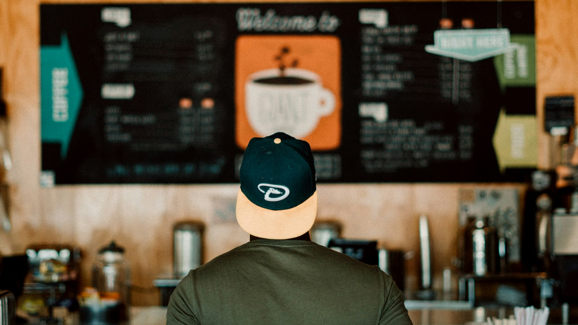 A person with a backwards baseball cap stands in front of coffee shop menu board, blurred and out of focused. 