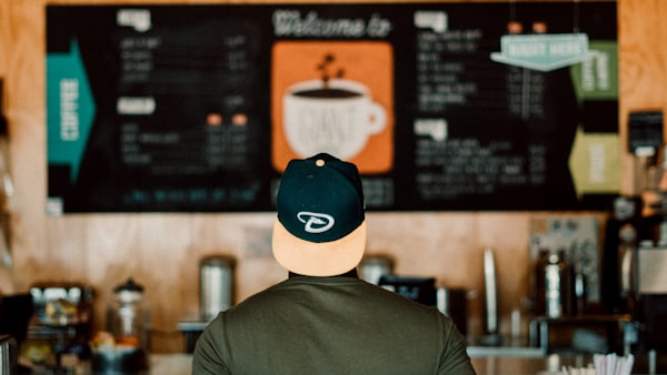 A person with a backwards baseball cap stands in front of coffee shop menu board, blurred and out of focused. 