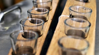 Several small empty glass cups are arranged on a wooden tray. The image is focused on the cups, with a blurred metallic surface in the background, suggesting a kitchen or bar setting.