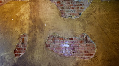 Exposed brickwork visible through patches of peeling plaster on a wall, revealing an irregular pattern. The surrounding plaster is a textured brown color, contrasting with the rough, red brick underneath.