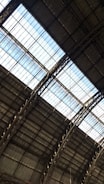 The image captures the interior view of a large industrial or warehouse building with a high ceiling. The roof structure is made up of metal beams and girders, with large glass panels letting in natural light. The overall architecture appears sturdy and functional.