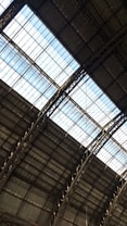 The image captures the interior view of a large industrial or warehouse building with a high ceiling. The roof structure is made up of metal beams and girders, with large glass panels letting in natural light. The overall architecture appears sturdy and functional.
