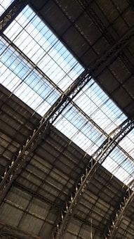 The image captures the interior view of a large industrial or warehouse building with a high ceiling. The roof structure is made up of metal beams and girders, with large glass panels letting in natural light. The overall architecture appears sturdy and functional.