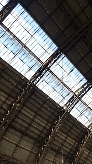 The image captures the interior view of a large industrial or warehouse building with a high ceiling. The roof structure is made up of metal beams and girders, with large glass panels letting in natural light. The overall architecture appears sturdy and functional.