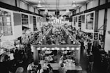 A bustling indoor market with multiple stalls, packed with people dining and shopping. The setting is industrial with exposed beams and ample lighting. Various food stalls and vendors line both sides of a central aisle, and patrons are seated at tables in the middle.