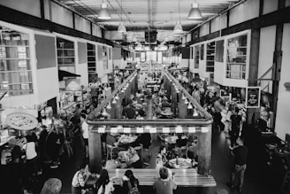A bustling indoor market with multiple stalls, packed with people dining and shopping. The setting is industrial with exposed beams and ample lighting. Various food stalls and vendors line both sides of a central aisle, and patrons are seated at tables in the middle.