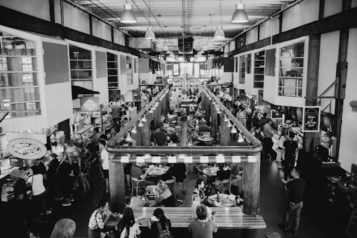 A bustling indoor market with multiple stalls, packed with people dining and shopping. The setting is industrial with exposed beams and ample lighting. Various food stalls and vendors line both sides of a central aisle, and patrons are seated at tables in the middle.
