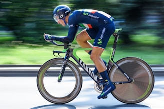 A professional cyclist in a blue and green uniform is speedily riding a time-trial bike with aerodynamic designs. The background is motion-blurred, emphasizing the sense of speed.