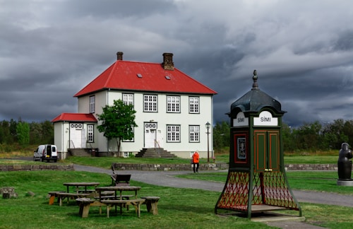 A traditional house with a white façade and a bright red roof is situated in a grassy area. In the foreground, there's an old-fashioned public phone booth with 'SÍMI' written on it. A person wearing an orange jacket is walking nearby, and a white van is parked to the left side. The sky is overcast with dark, dramatic clouds.