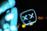 A neon light art piece displays the outline of a face with X-shaped eyes and a stitched mouth pattern. The image is dark, emphasizing the bright cyan and yellow lights, which form abstract shapes.