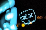 A neon light art piece displays the outline of a face with X-shaped eyes and a stitched mouth pattern. The image is dark, emphasizing the bright cyan and yellow lights, which form abstract shapes.