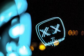 A neon light art piece displays the outline of a face with X-shaped eyes and a stitched mouth pattern. The image is dark, emphasizing the bright cyan and yellow lights, which form abstract shapes.