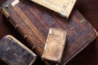 A collection of old, ornate books with richly carved and aged leather covers, featuring intricate designs and embossments. The books are stacked on a wooden surface, displaying their worn and historic appearance.