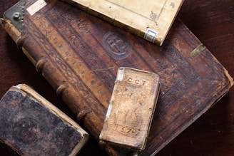 A collection of old, ornate books with richly carved and aged leather covers, featuring intricate designs and embossments. The books are stacked on a wooden surface, displaying their worn and historic appearance.